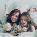 Laughing happy mother and cute daughter watching funny cartoon movie on TV while lying on bed at home in morning have fun at cozy bedroom